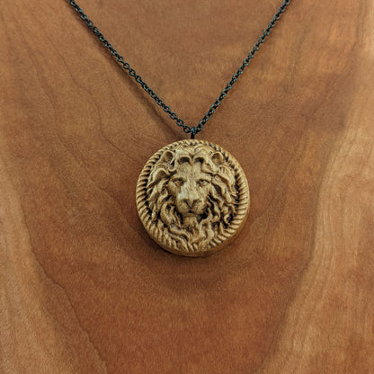 Wooden necklace pendant carved in the shape of a stern lion face surrounded by a rope border. Hanging from a black stainless steel chain against a cherry wood background.