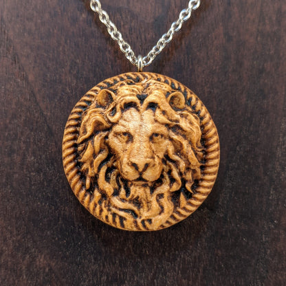 Wooden necklace pendant carved in the shape of a stern lion face surrounded by a rope border. Hanging from a silver stainless steel chain against a dark walnut wood background.