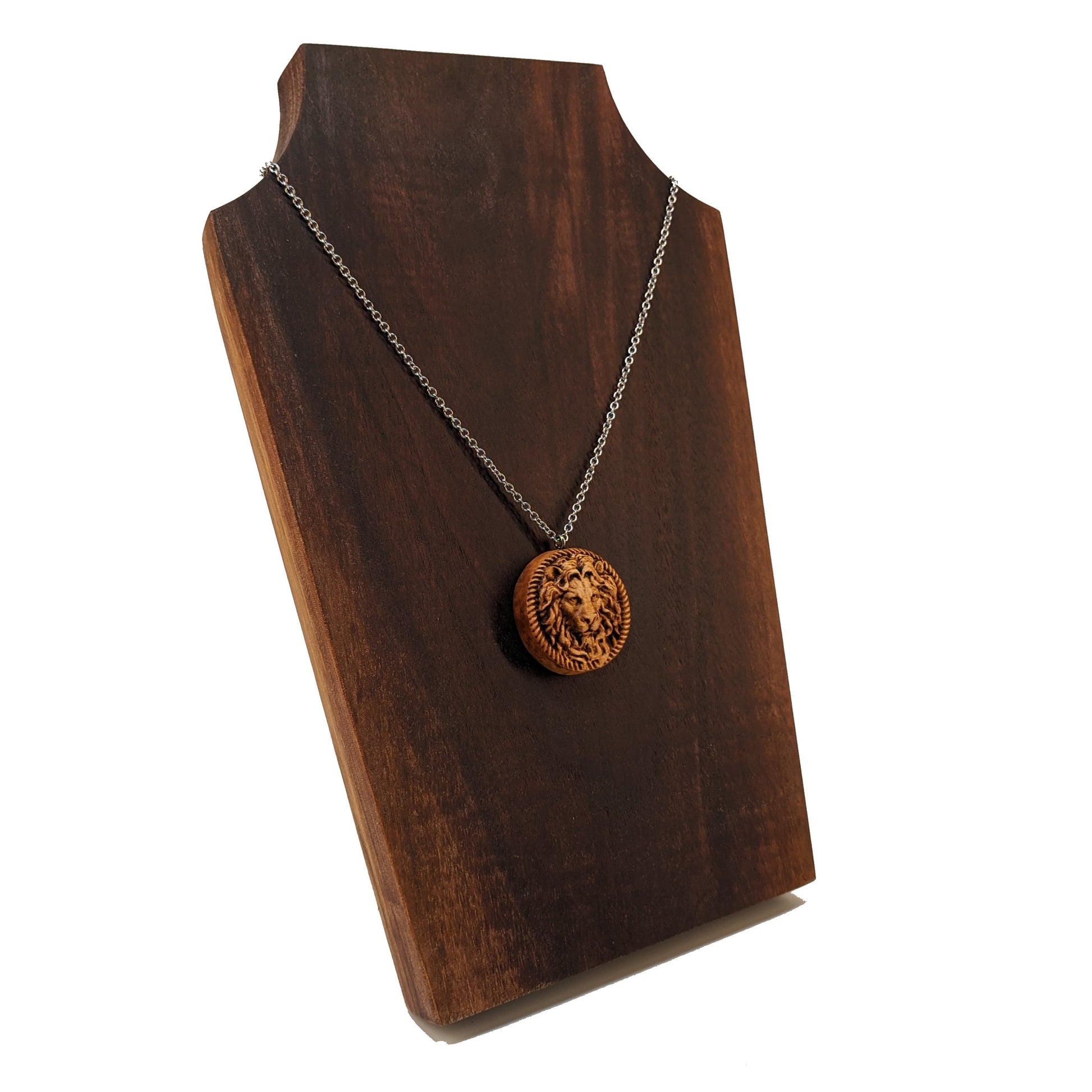 Wooden necklace pendant carved in the shape of a stern lion face surrounded by a rope border. Hanging from a silver stainless steel chain against a dark walnut wood background.