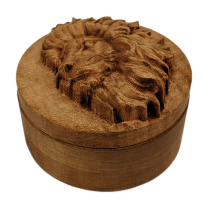 Round wooden box 3D carved with a side profile of a lions face. It has a serious look in its brow with a closed mouth facing to the left with a large bushy mane surrounding its face. Made from hard maple wood stained brown against a white background.