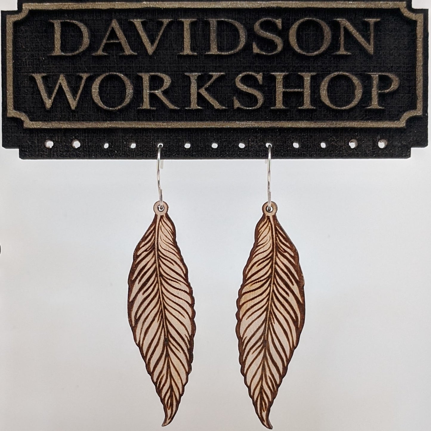 Pair of wooden earrings with silver stainless steel hooks. They are light natural finished, long realistically engraved feathers. Made from birch wood hanging from a model Davidson Workshop sign against a white background. (Close up View)