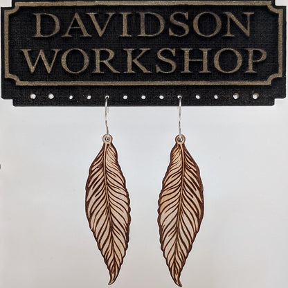 Pair of wooden earrings with silver stainless steel hooks. They are light natural finished, long realistically engraved feathers. Made from birch wood hanging from a model Davidson Workshop sign against a white background. (Close up View)