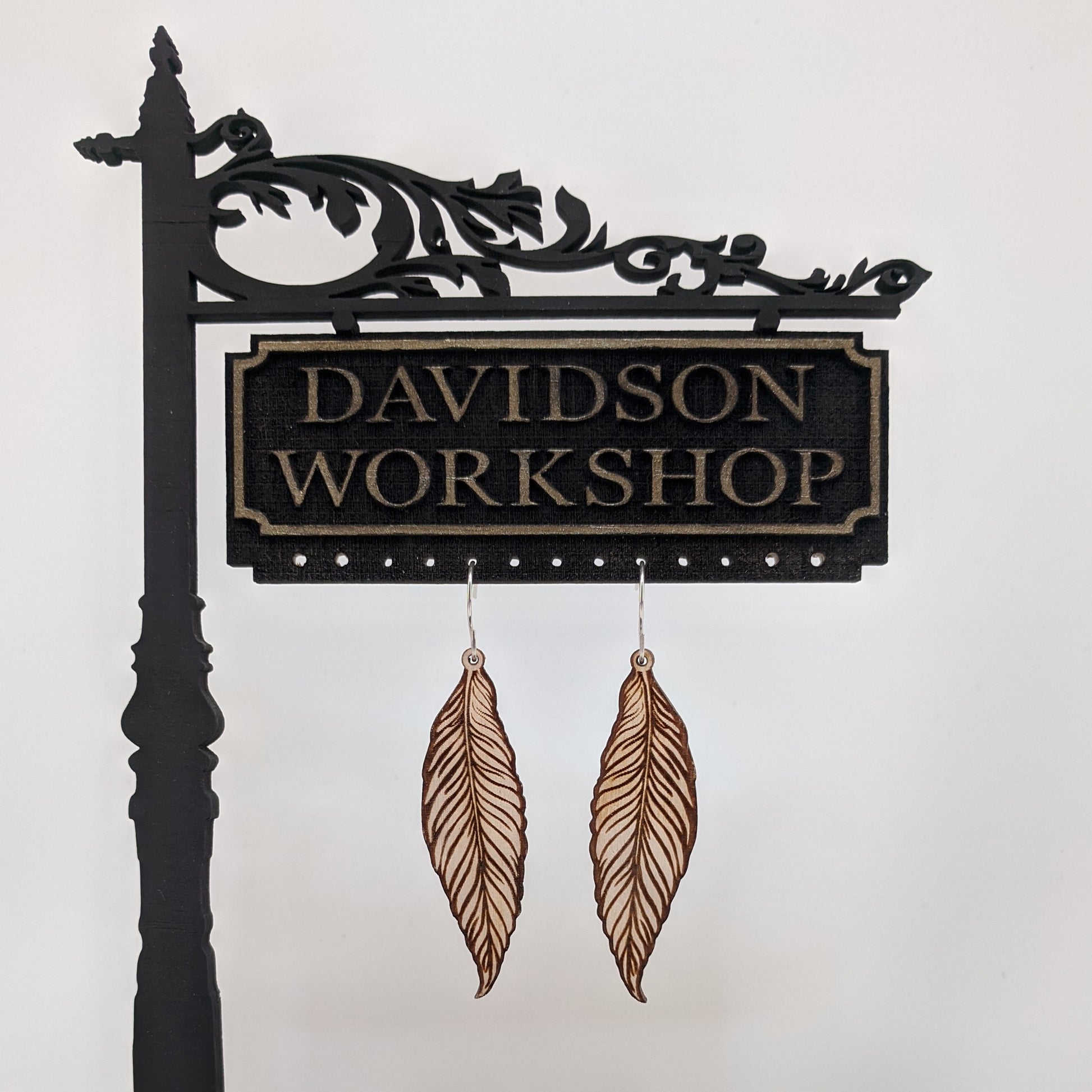 Pair of wooden earrings with silver stainless steel hooks. They are pale natural finished, long and slim realistic engraved feathers. Made from birch wood hanging from a model Davidson Workshop sign against a white background. 