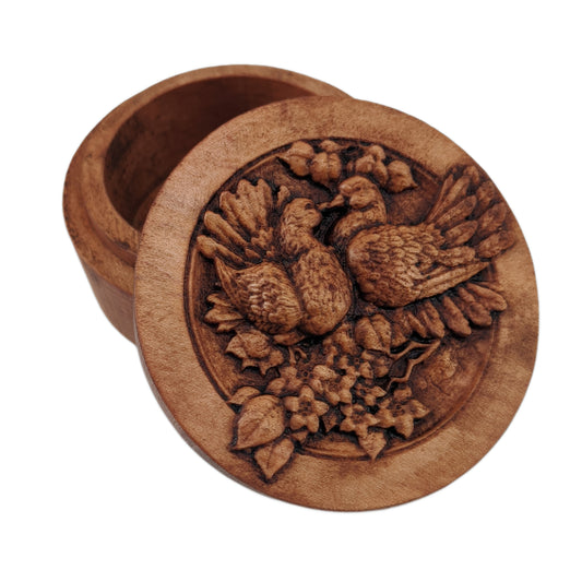 Round wooden box 3D carved with two lovebirds standing on a branch blooming with flowers, looking at each other with their beaks touching and feathers fluffed up. Made from hard maple wood stained brown against a white background.