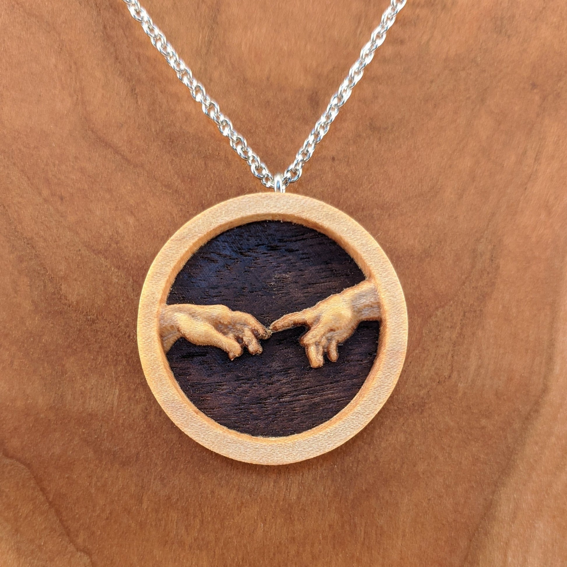 Round wooden necklace pendant carved in the shape of 2 hands reaching out to touch each other. The hands are carved from light maple wood overlayed on a dark walnut base. Hanging from a silver stainless steel chain against a cherry wood background.