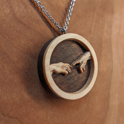 Round wooden necklace pendant carved in the shape of 2 hands reaching out to touch each other. The hands are carved from light maple wood overlayed on a dark walnut base. Hanging from a silver stainless steel chain against a cherry wood background.