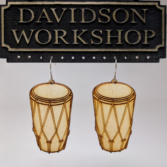 Pair of wooden earrings with silver stainless steel hooks. They are pale natural finished bongo drums with engraved rope woven across the side. Made from birch wood hanging from a model Davidson Workshop sign against a white background(Close up View)