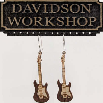 Pair of wooden earrings with silver stainless steel hooks. They are two toned light and dark brown Stratocaster style electric guitars.  Made from birch wood hanging from a model Davidson Workshop sign against a white background. (Close up View)