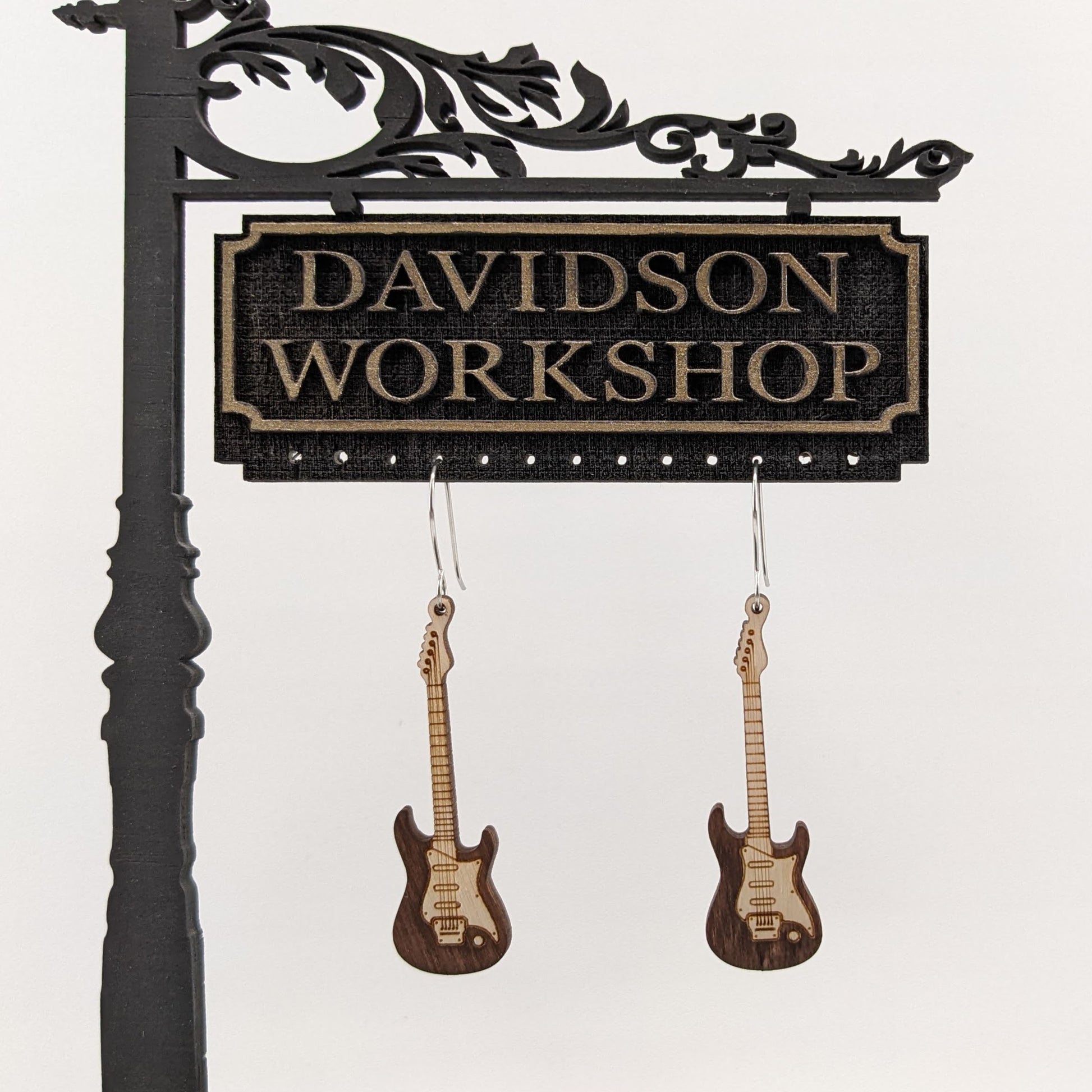 Pair of wooden earrings with silver stainless steel hooks. They are two toned light and dark brown Stratocaster style electric guitars.  Made from birch wood hanging from a model Davidson Workshop sign against a white background.