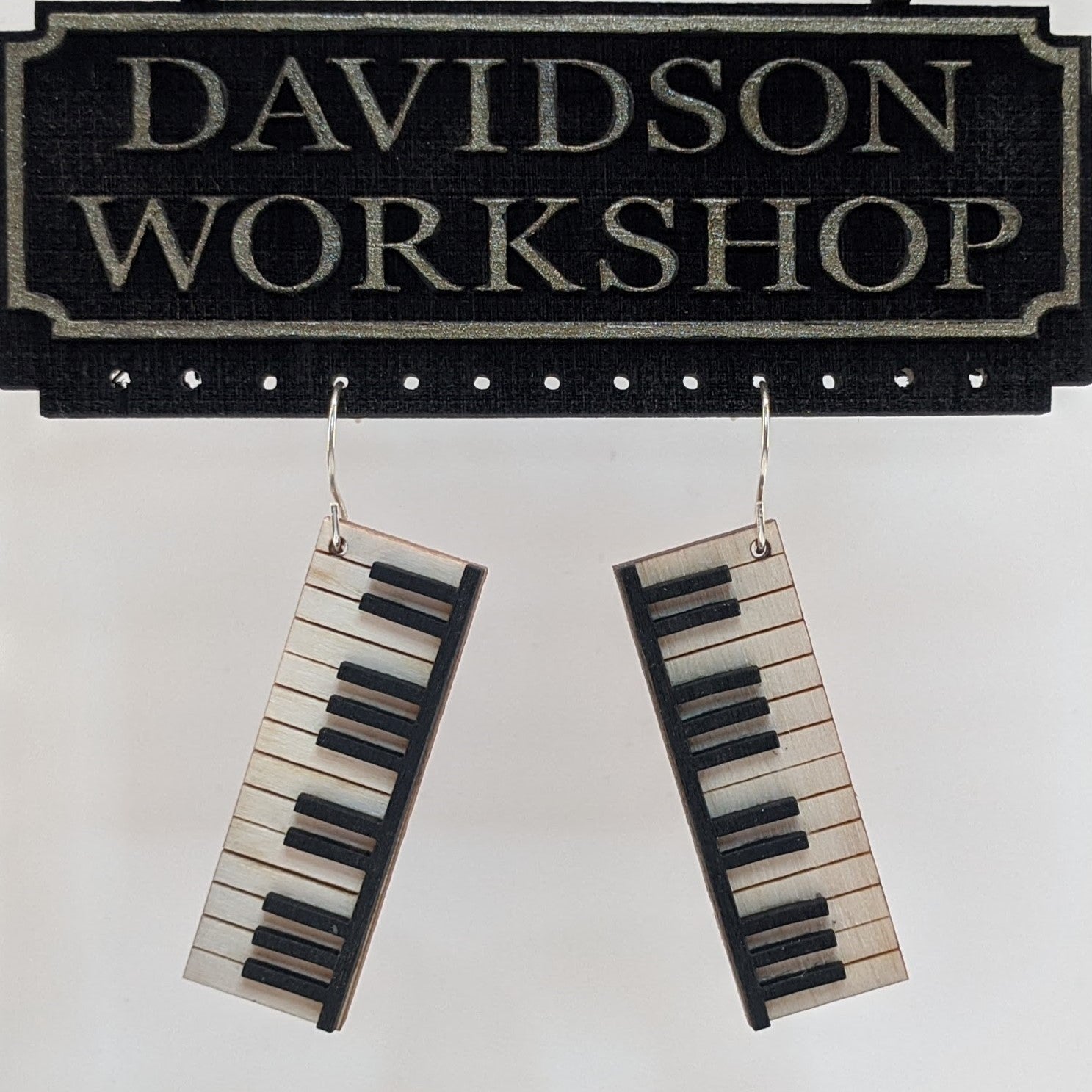 Pair of wooden earrings with silver stainless steel hooks. They are pale natural finished miniature keyboards with painted black keys. Made from birch wood hanging from a model Davidson Workshop sign against a white background. (Close up View)