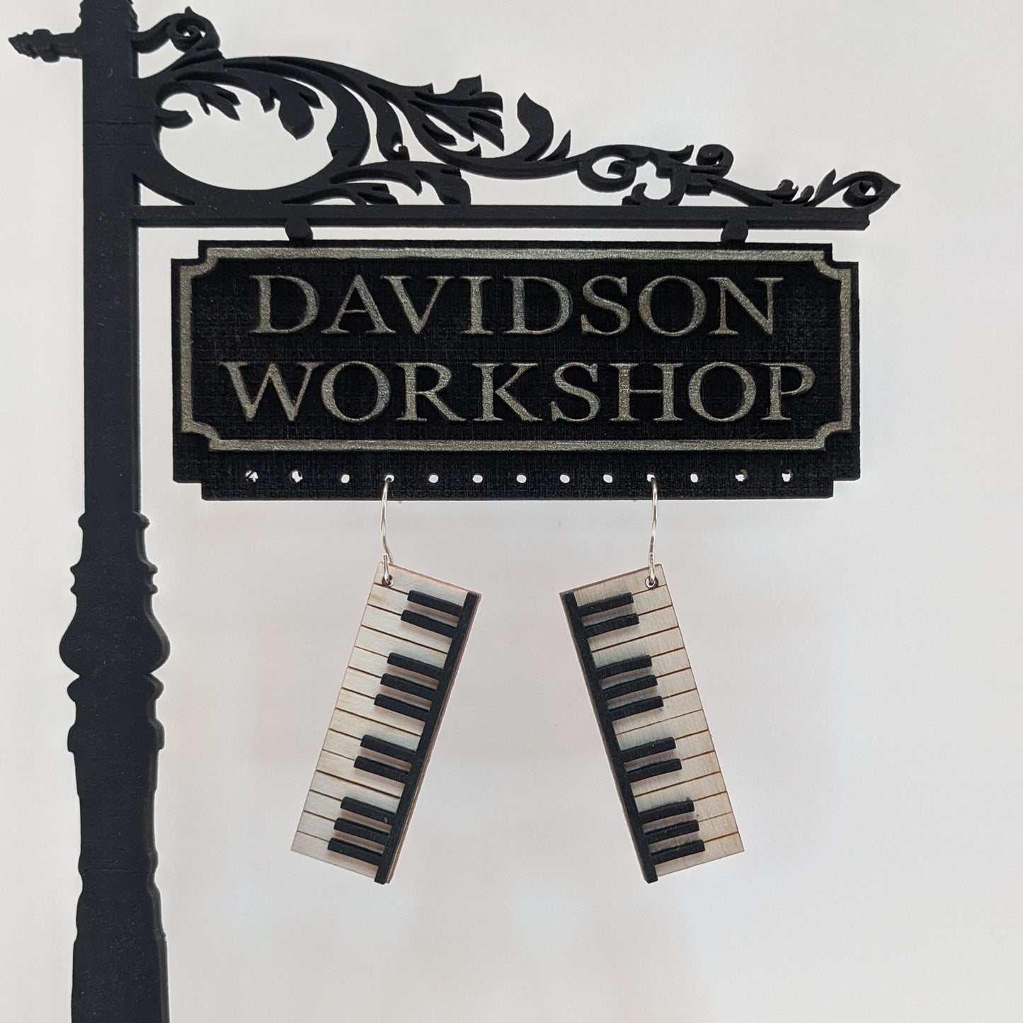 Pair of wooden earrings with silver stainless steel hooks. They are pale natural finished miniature keyboards with painted black keys. Made from birch wood hanging from a model Davidson Workshop sign against a white background.