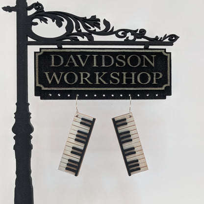 Pair of wooden earrings with silver stainless steel hooks. They are pale natural finished miniature keyboards with painted black keys. Made from birch wood hanging from a model Davidson Workshop sign against a white background.