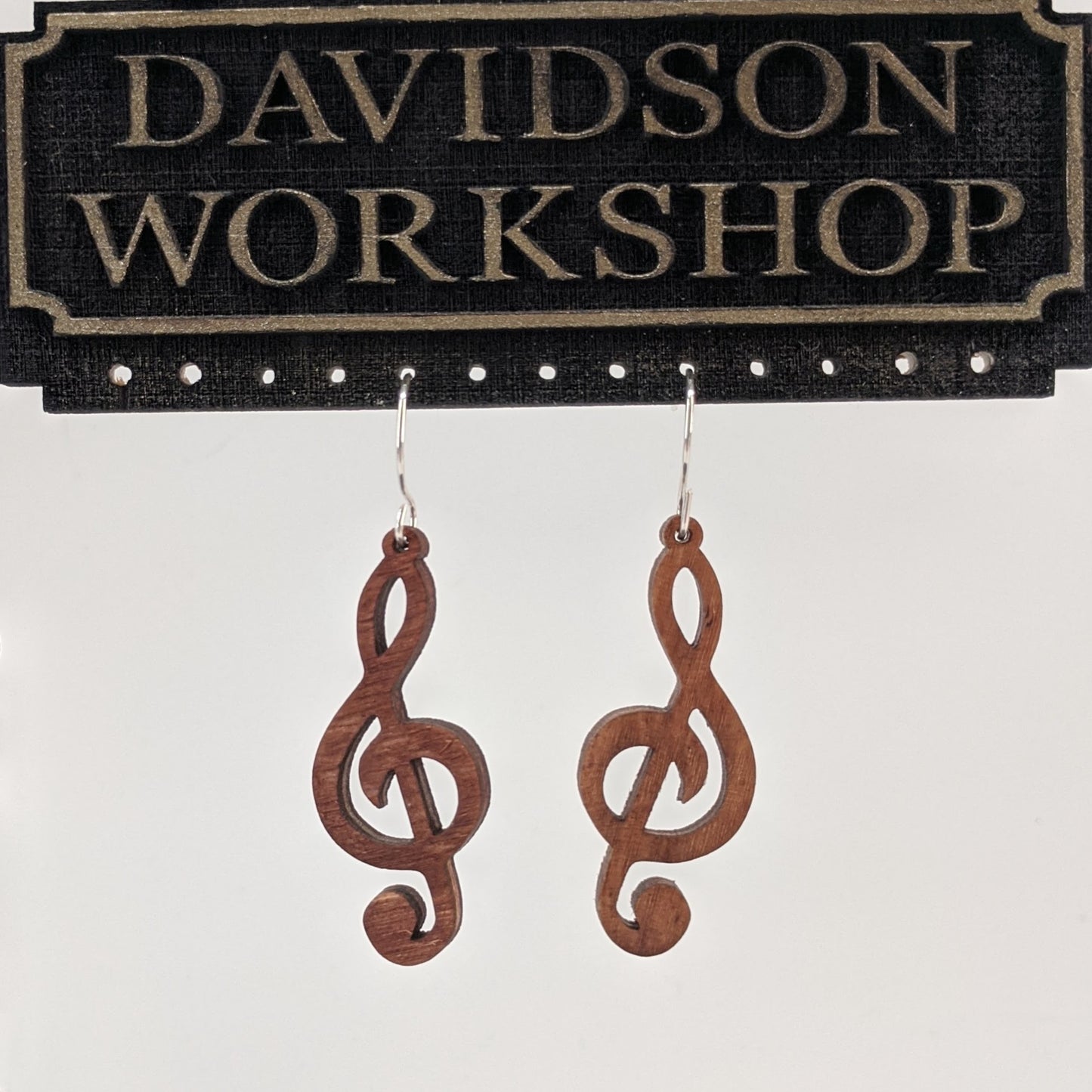 Pair of wooden earrings with silver stainless steel hooks. They are brown cutouts of the musical notes treble clef. Made from birch wood hanging from a model Davidson Workshop sign against a white background. (Close up View)