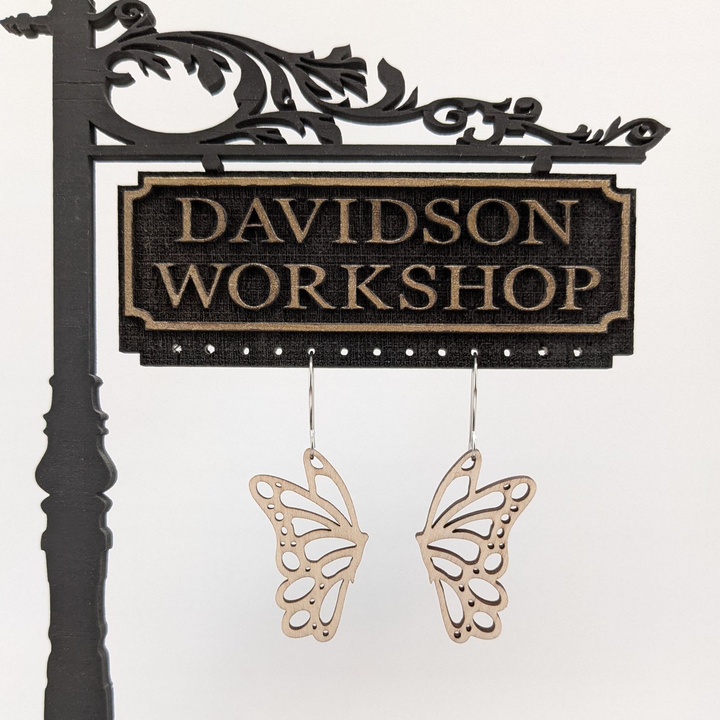 Pair of wooden earrings with silver stainless steel hooks. They are pale natural finished cutouts of side profile butterflies. Made from birch wood hanging from a model Davidson Workshop sign against a white background. 