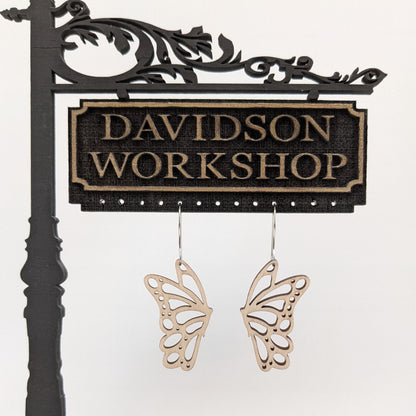 Pair of wooden earrings with silver stainless steel hooks. They are pale natural finished cutouts of side profile butterflies. Made from birch wood hanging from a model Davidson Workshop sign against a white background. 