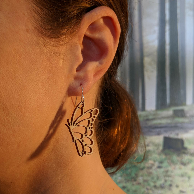 Pair of wooden earrings with silver stainless steel hooks. They are pale natural finished cutouts of side profile butterflies. Made from birch wood, being modeled by a women in front of a nature scene.