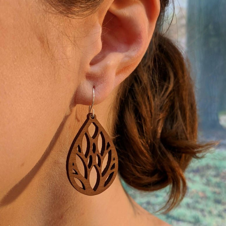 Pair of wooden earrings with silver stainless steel hooks. They are brown teardrop shaped. With multiple eye and almond shaped cutouts within. Made from birch wood, being modeled by a women in front of a nature scene.
