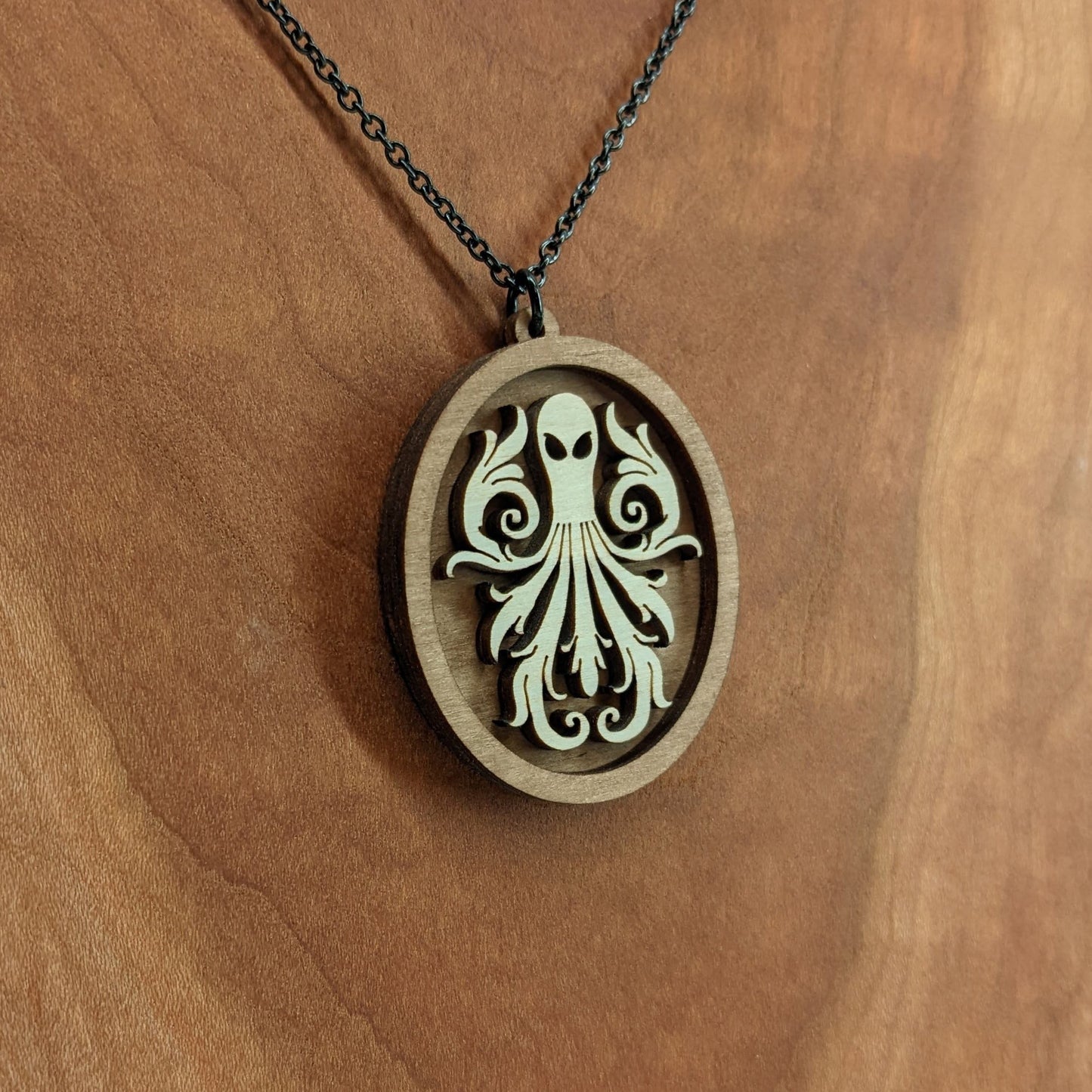 Oval wooden octopus pendant necklace made for contrasting layers of light and dark birch wood. The Octopus is light in color overlayed onto a darker base. Hanging from a black stainless steel chain against a cherry wood background.