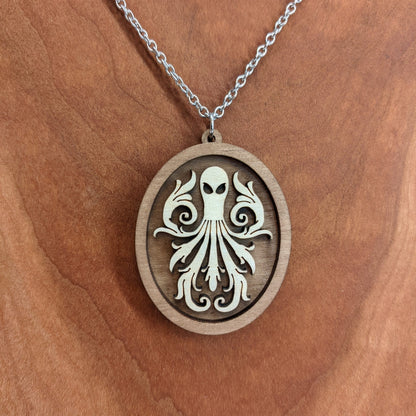 Oval wooden octopus pendant necklace made of contrasting layers of light and dark birch wood. The Octopus is light in color overlayed onto a darker base. Hanging from a silver stainless steel chain against a cherry wood background.