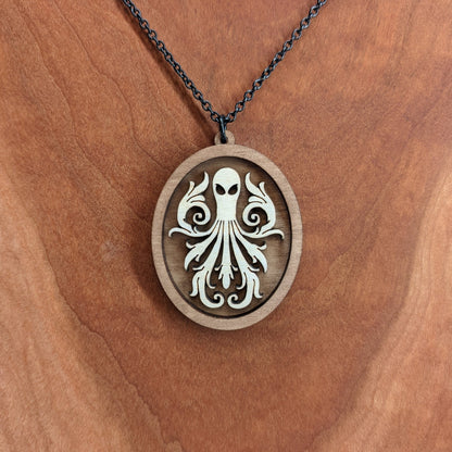 Oval wooden octopus pendant necklace made for contrasting layers of light and dark birch wood. The Octopus is light in color overlayed onto a darker base. Hanging from a black stainless steel chain against a cherry wood background.