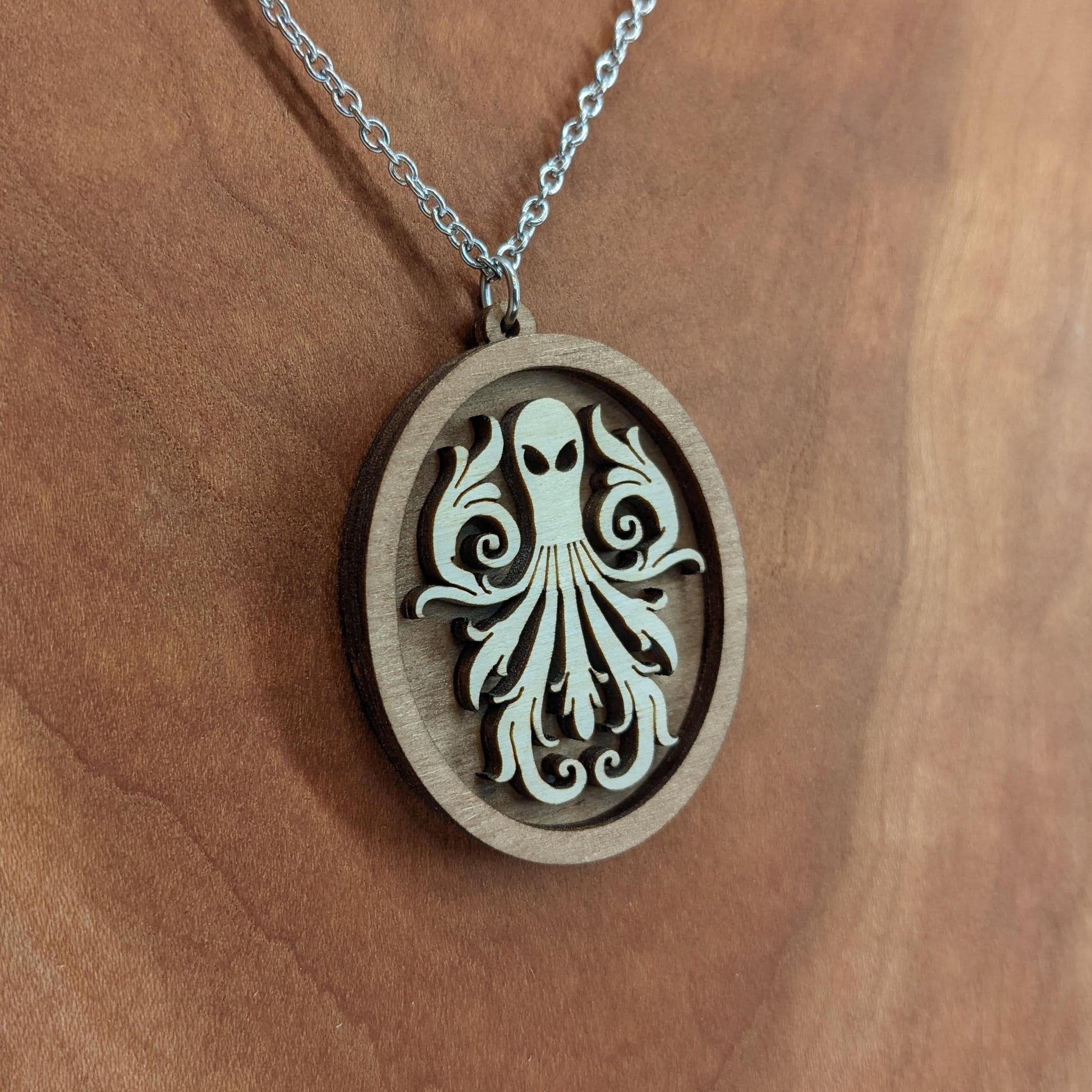 Oval wooden octopus pendant necklace made of contrasting layers of light and dark birch wood. The Octopus is light in color overlayed onto a darker base. Hanging from a silver stainless steel chain against a cherry wood background.