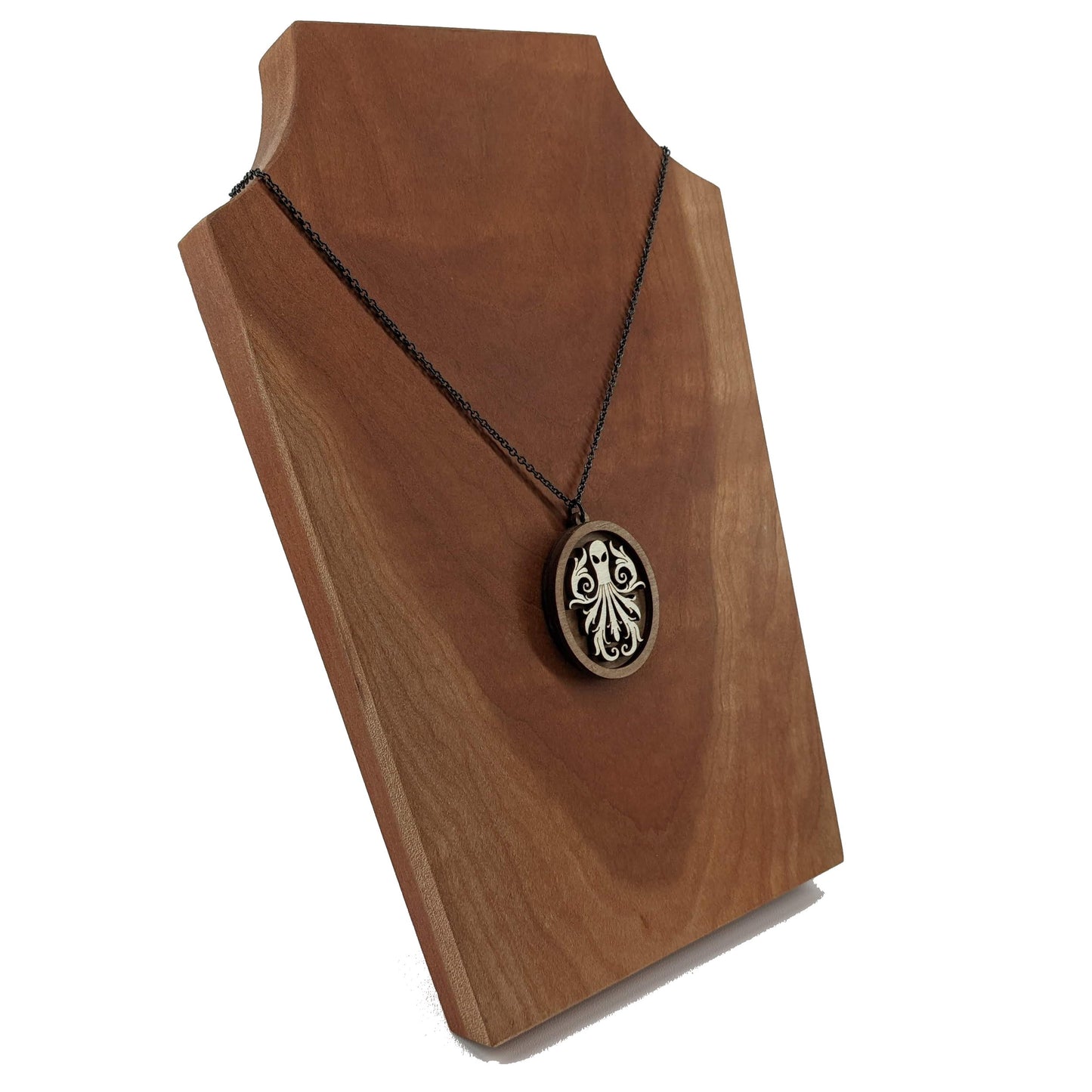 Oval wooden octopus pendant necklace made for contrasting layers of light and dark birch wood. The Octopus is light in color overlayed onto a darker base. Hanging from a black stainless steel chain against a cherry wood background.