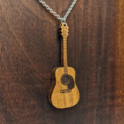 Wooden necklace pendant created in the shape of a miniature acoustic guitar . Made from maple wood with an antique finish and hanging from a black stainless steel chain against a dark walnut background.