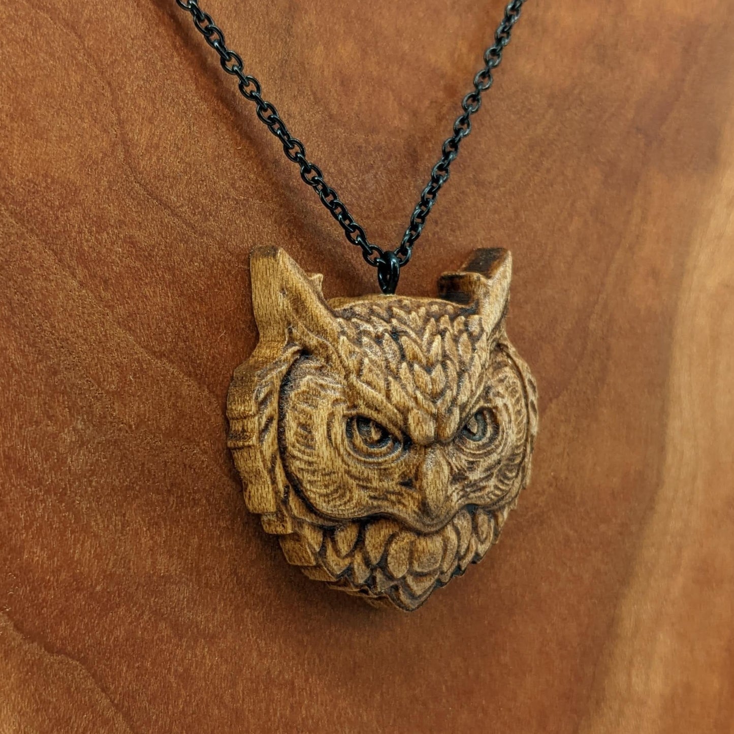 Wooden necklace pendant carved in the shaped of an owls head. Made from hard maple wood. Hanging from a black stainless steel chain against a cherry wood background.