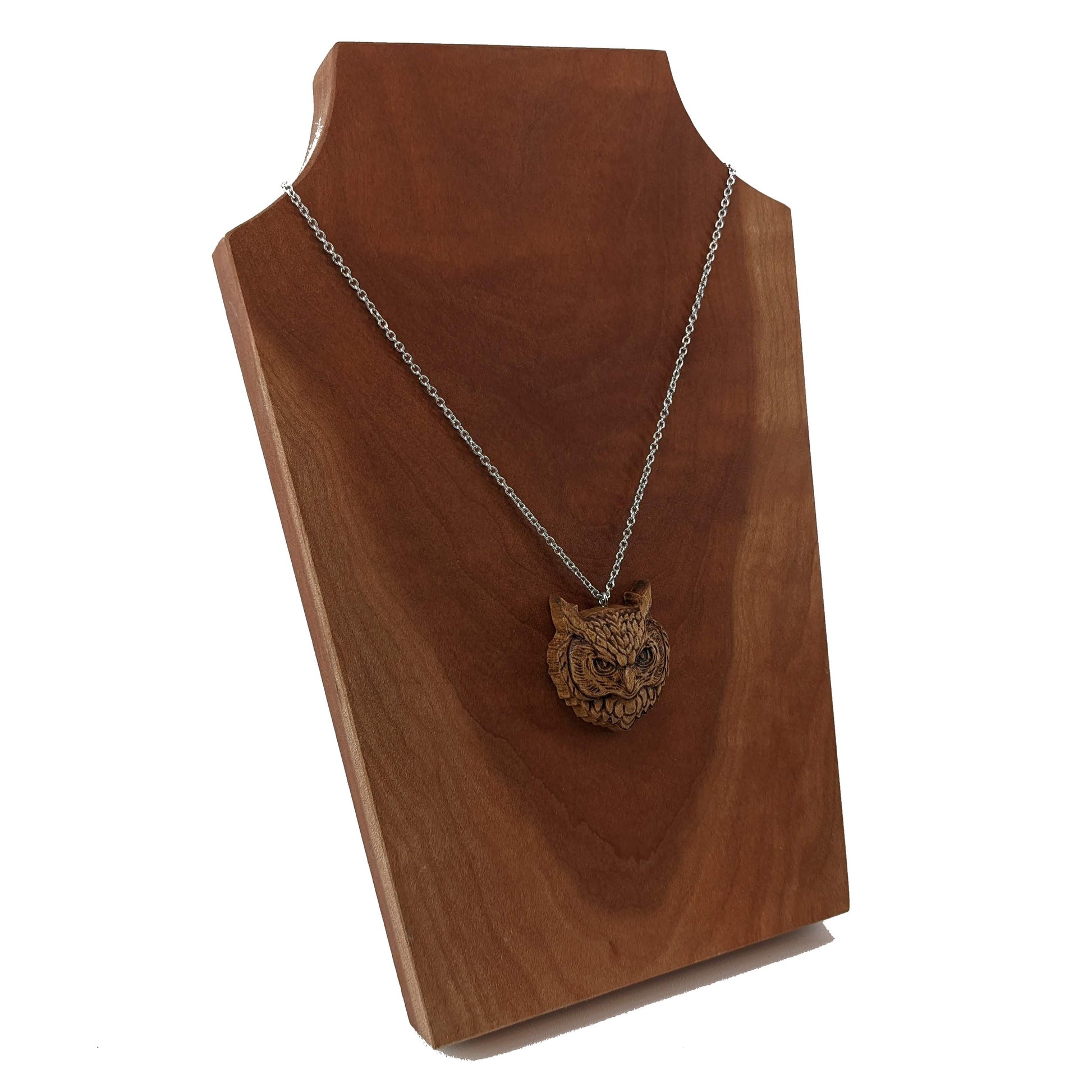 Wooden necklace pendant carved in the shaped of an owls head. Made from hard maple wood. Hanging from a silver stainless steel chain against a cherry wood background.