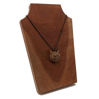 Wooden necklace pendant carved in the shaped of an owls head. Made from hard maple wood. Hanging from a black stainless steel chain against a cherry wood background.