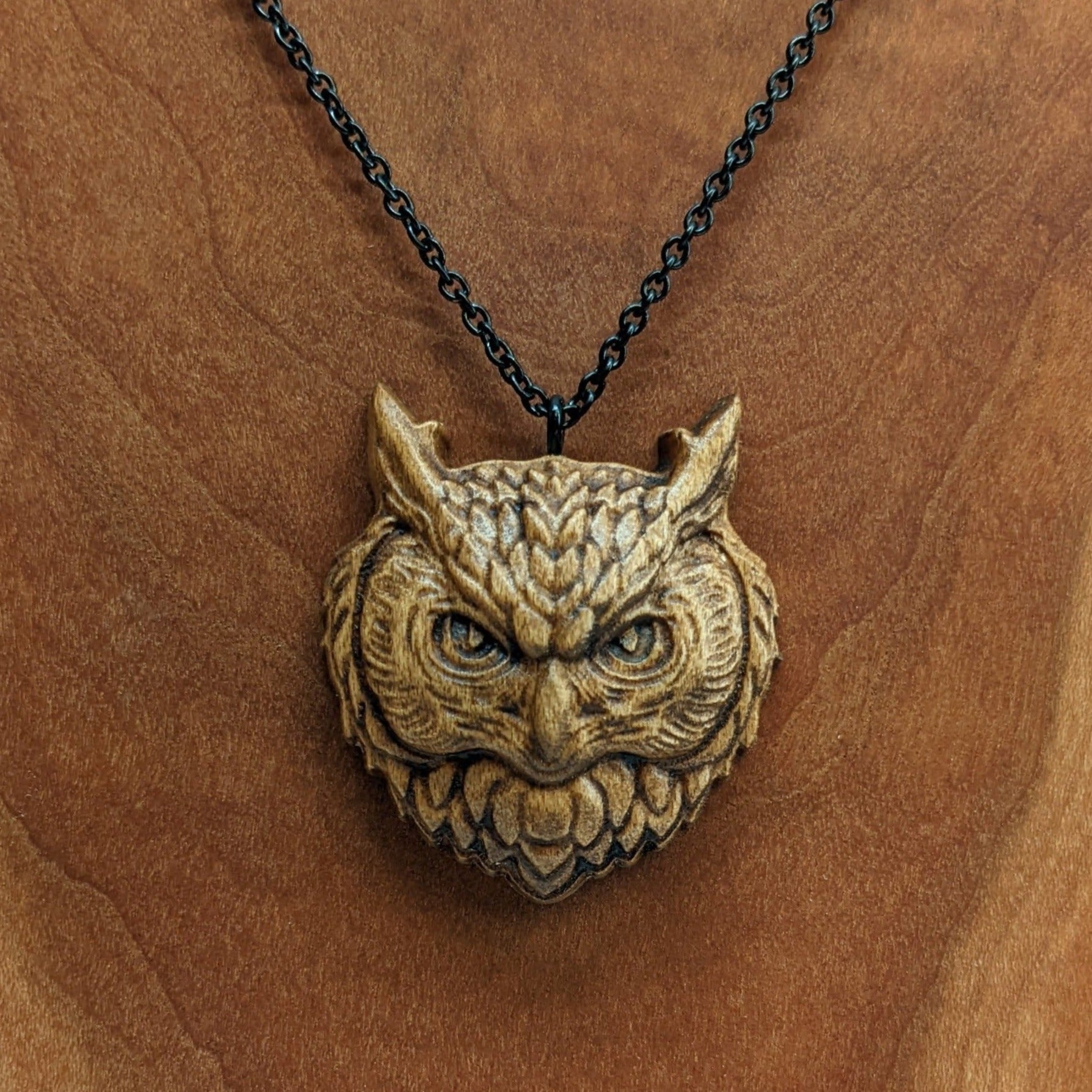 Wooden necklace pendant carved in the shaped of an owls head. Made from hard maple wood. Hanging from a black stainless steel chain against a cherry wood background.