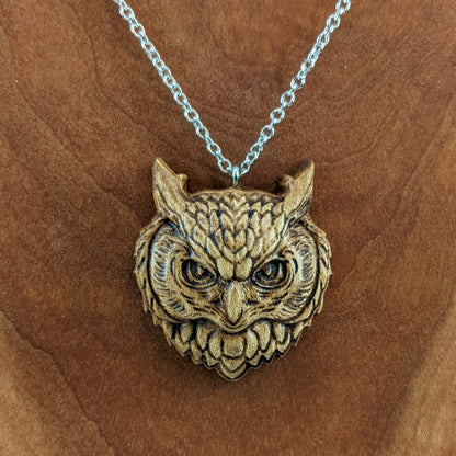 Wooden necklace pendant carved in the shaped of an owls head. Made from hard maple wood. Hanging from a silver stainless steel chain against a cherry wood background.