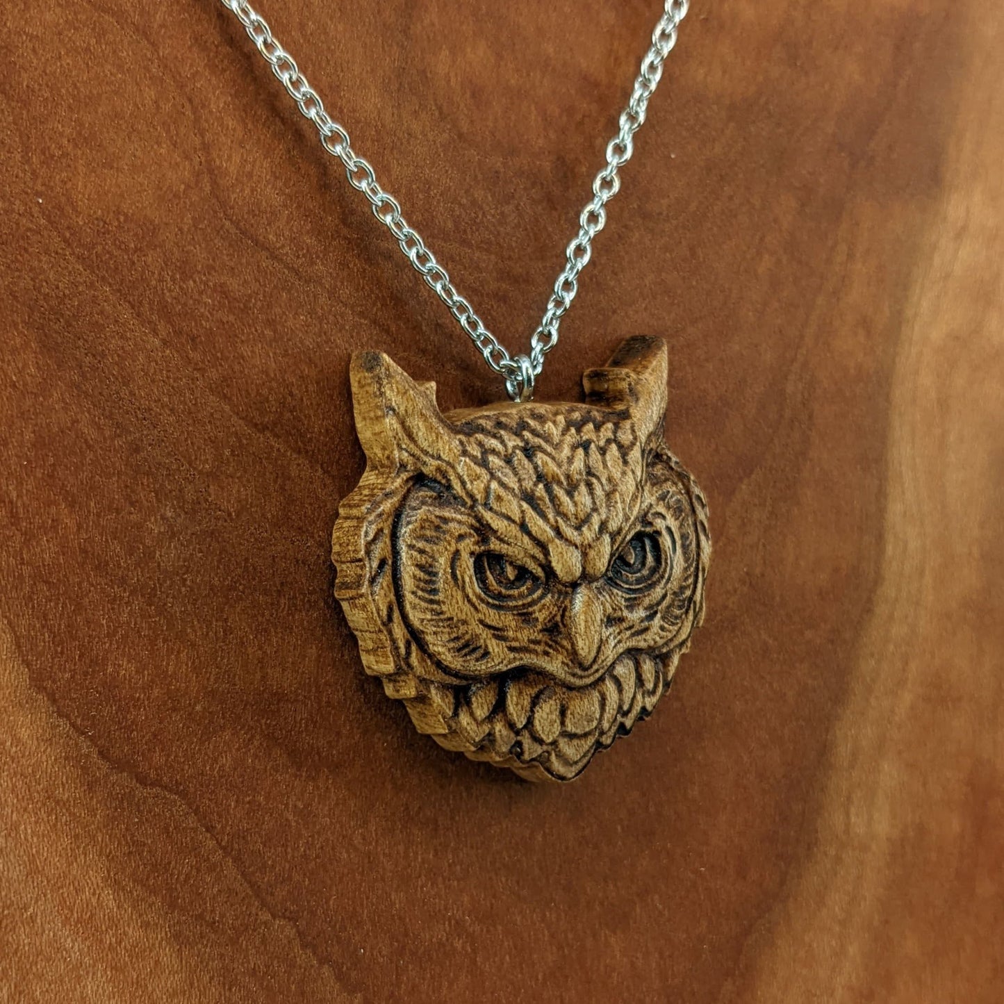 Wooden necklace pendant carved in the shaped of an owls head. Made from hard maple wood. Hanging from a silver stainless steel chain against a cherry wood background.