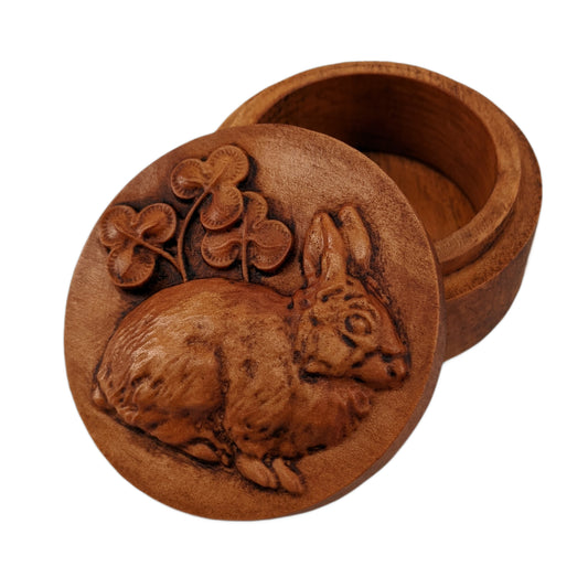 Round wooden box 3D carved with a fluffy lucky rabbit sitting, turned to the right, with its ears perked up and  clovers growing up from behind it. Made from hard maple wood stained brown against a white background.