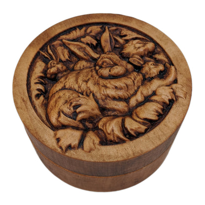 Round wooden box 3D carved with a family of rabbits together in their nest. Mother rabbit has bushy fur, tall ears and  heads of three baby rabbits peaking out from behind her. Made from hard maple wood stained brown against a white background.