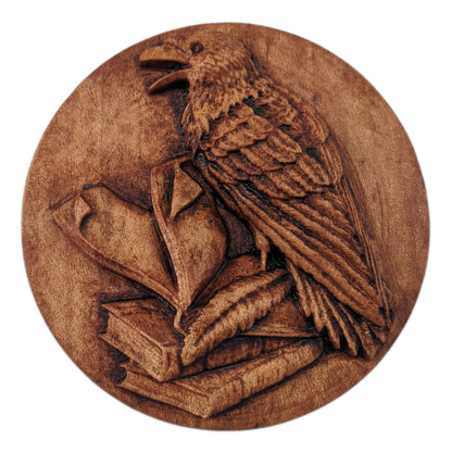 Round wooden box 3D carved with a raven standing three books with a quill at its feet, the top book open and pages  turning. The raven has an open beak and intricate layered feathers Made from hard maple wood stained brown against a white background.