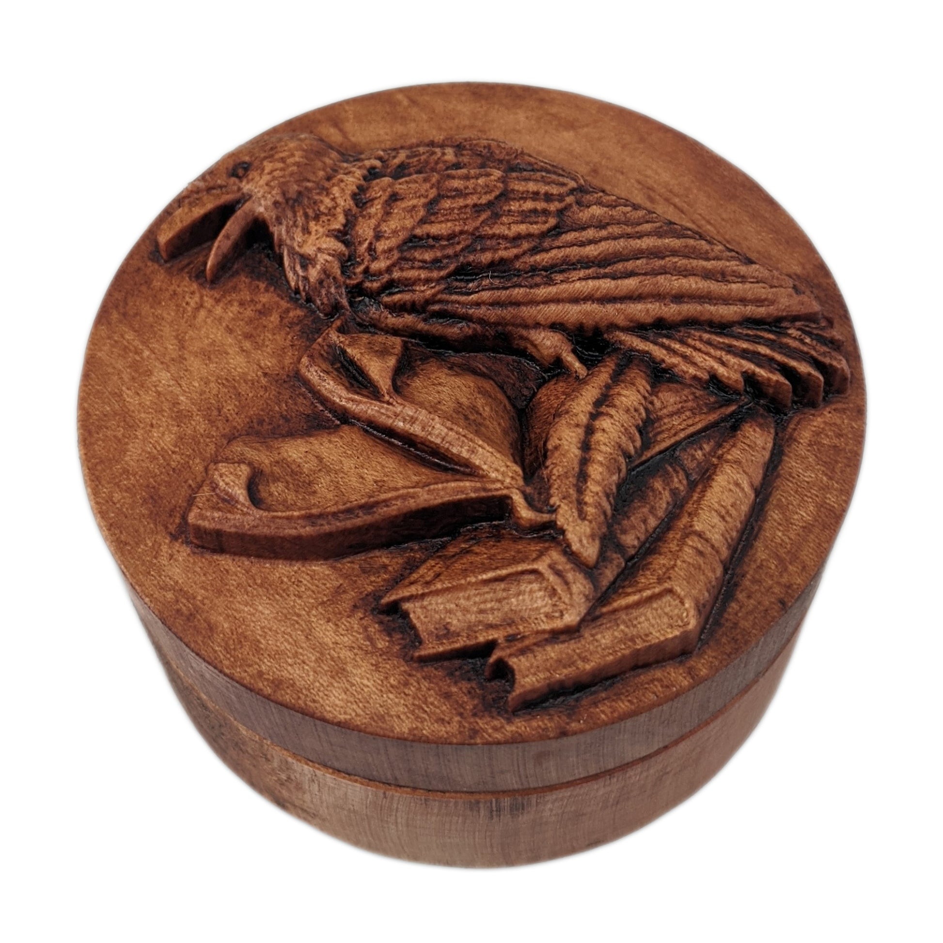 Round wooden box 3D carved with a raven standing three books with a quill at its feet, the top book open and pages  turning. The raven has an open beak and intricate layered feathers Made from hard maple wood stained brown against a white background.