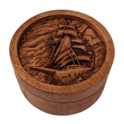 Round wooden box 3D carved with a large sailboat on the sea. It has multiple sails all pulled taught with ropes hanging all around. The background is a cloudy sky and rolling waves. Made from hard maple wood stained brown against a white background.