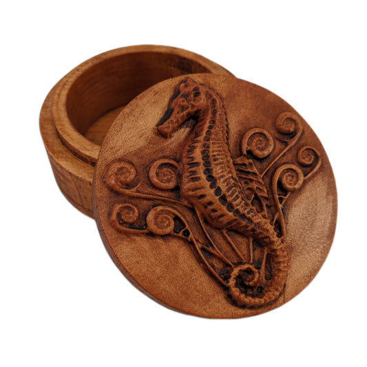 Round wooden box 3D carved with a seahorse turned to the left with rippled texture down its body and a curled tail. It has thin plants curled at the top surrounding it.  Made from hard maple wood stained brown against a white background.