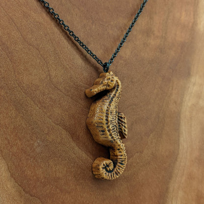 Wooden pendant necklace carved in the shape of sea horse. Made from hard maple and hanging from a black stainless steel chain against a white background.