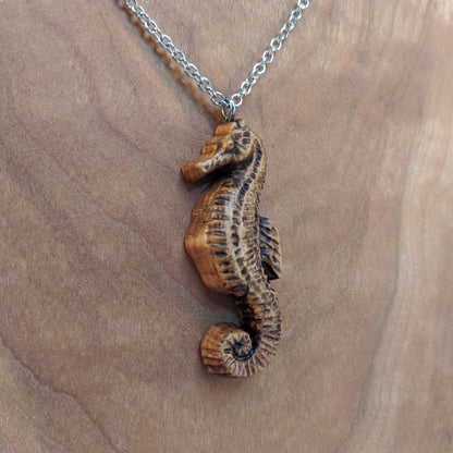 Wooden pendant necklace carved in the shape of sea horse. Made from hard maple and hanging from a silver stainless steel chain against a cherry wood background.