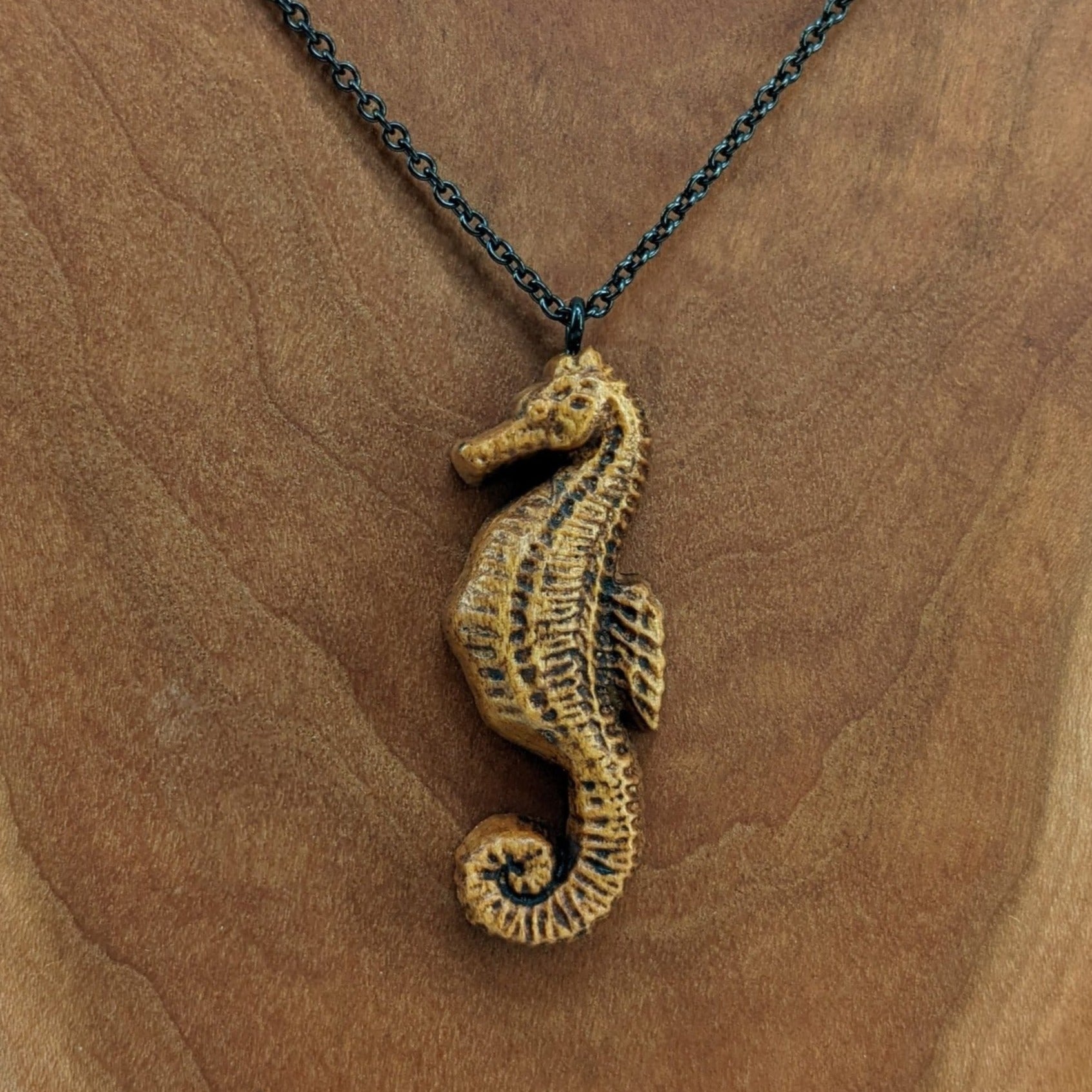 Wooden pendant necklace carved in the shape of sea horse. Made from hard maple and hanging from a black stainless steel chain against a white background.