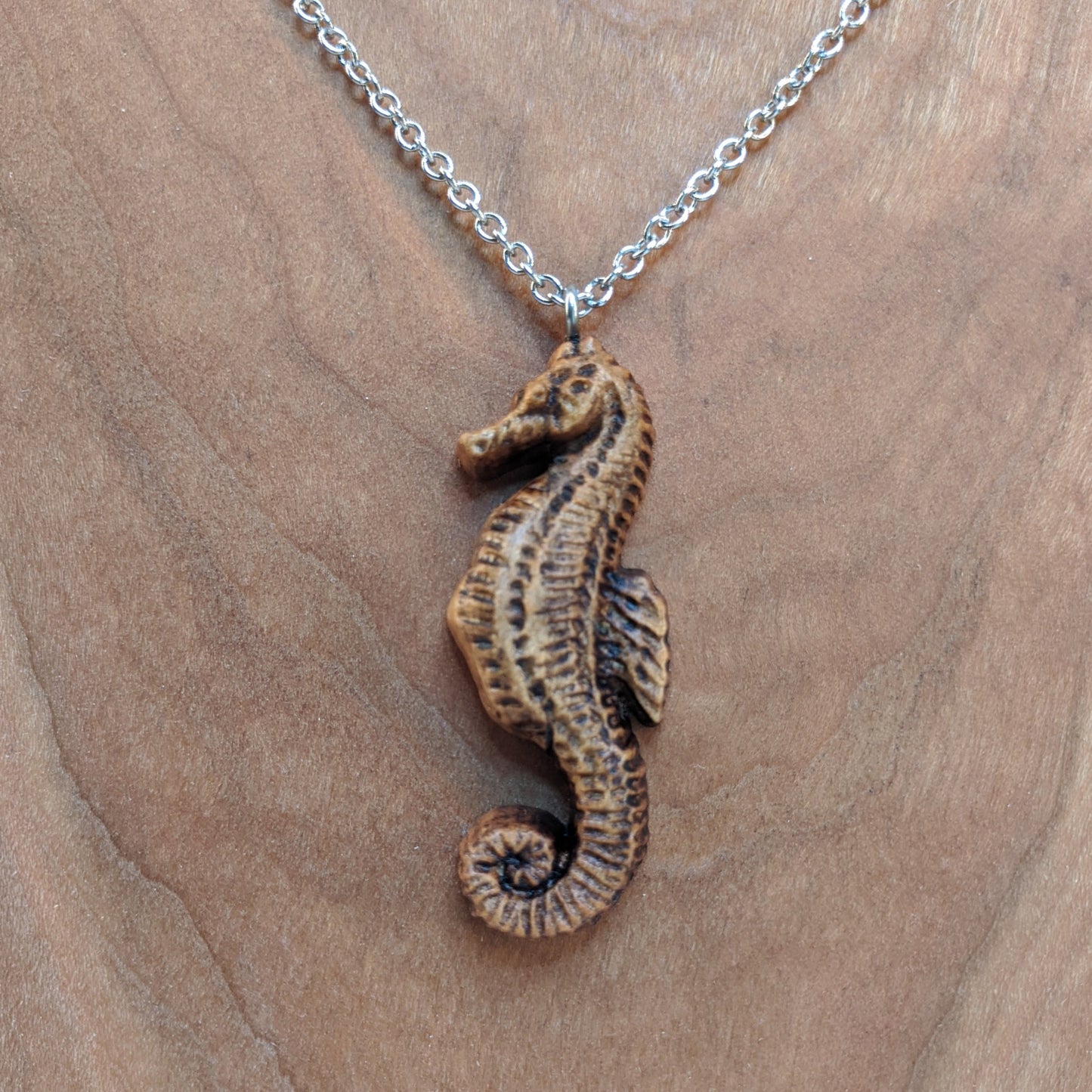 Wooden pendant necklace carved in the shape of sea horse. Made from hard maple and hanging from a silver stainless steel chain against a cherry wood background.