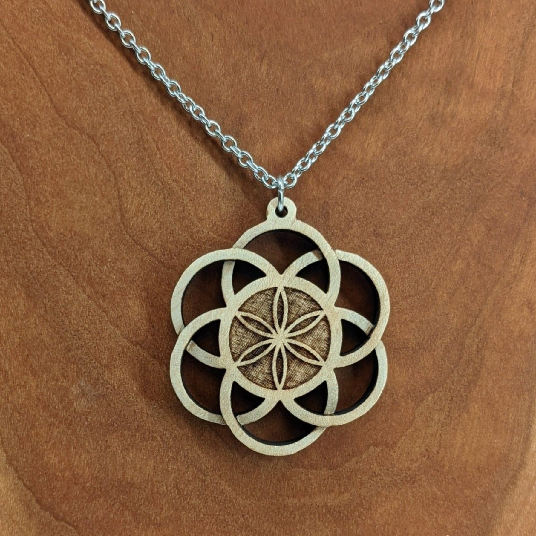 Wooden necklace pendant inspired by a flower of life including 6 interlinking circles creating a flower in the center. Carved from hard maple wood. Hanging from a black stainless steel chain against a cherry wood background.