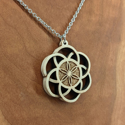 Wooden necklace pendant inspired by a flower of life including 6 interlinking circles creating a flower in the center. Carved from hard maple wood. Hanging from a black stainless steel chain against a cherry wood background.