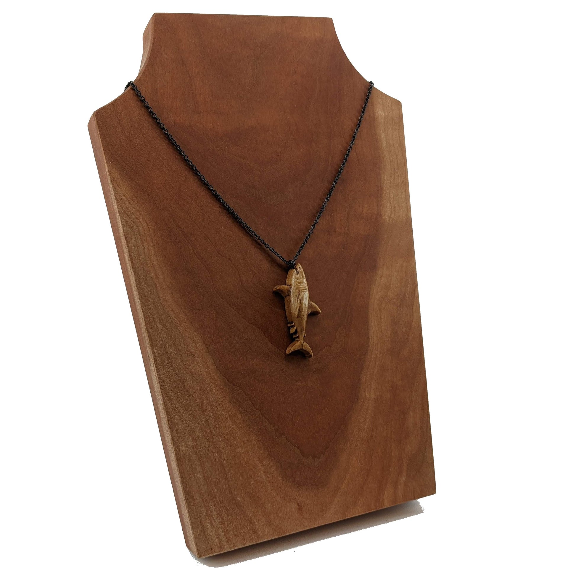 Wooden pendant necklace carved in the shape of great white shark. Made from hard maple wood. The wood shark hangs from its mouth on a black stainless steel chain against a cherry wood background.