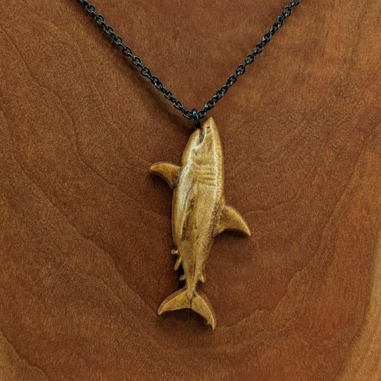 Wooden pendant necklace carved in the shape of great white shark. Made from hard maple wood. The wood shark hangs from its mouth on a black stainless steel chain against a cherry wood background.