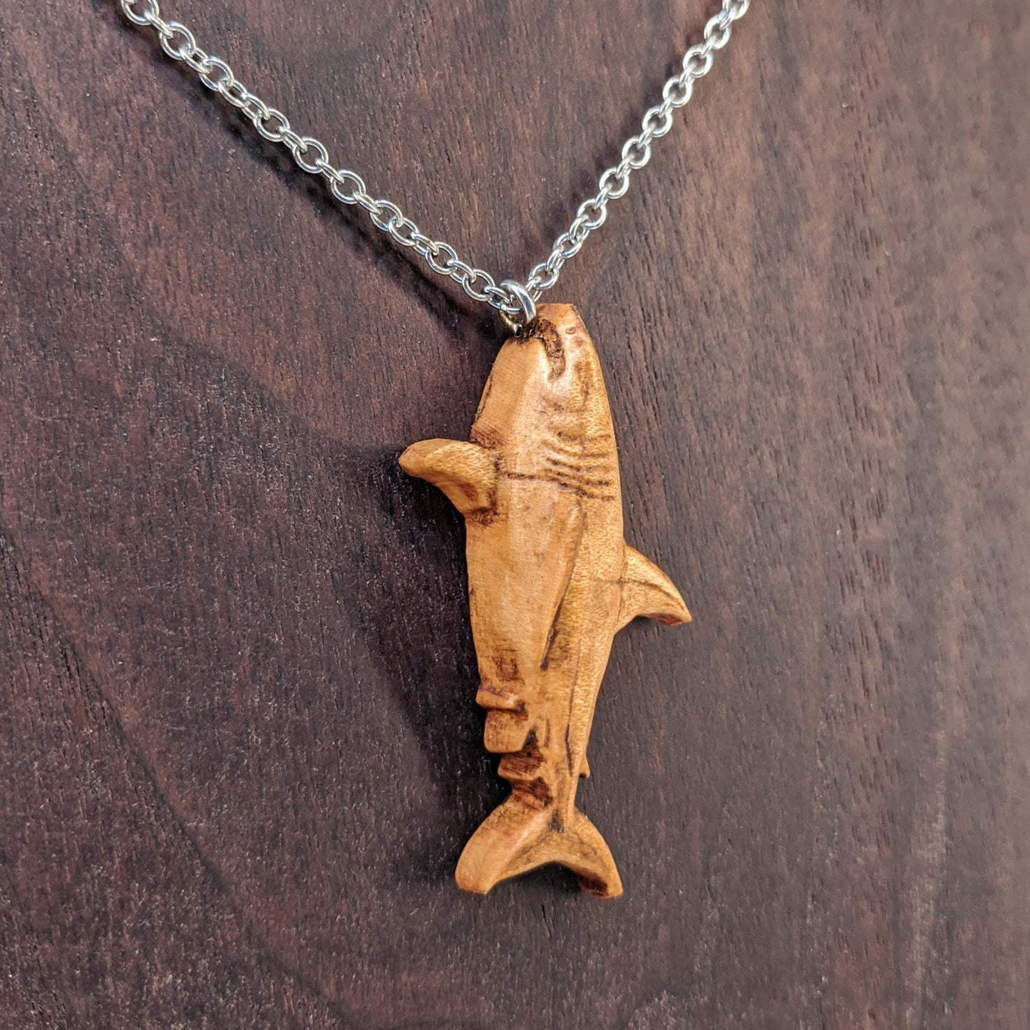 Wooden pendant necklace carved in the shape of great white shark. Made from hard maple wood. The wood shark hangs from its mouth on a black stainless steel chain against a dark walnut background.