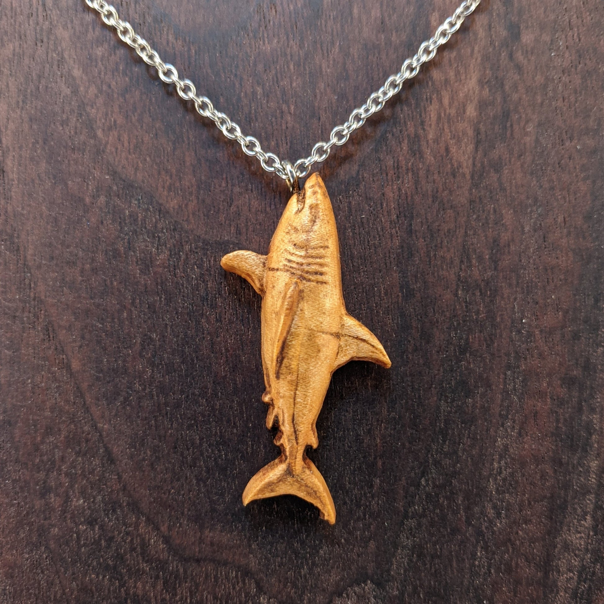 Wooden pendant necklace carved in the shape of great white shark. Made from hard maple wood. The wood shark hangs from its mouth on a black stainless steel chain against a dark walnut background.