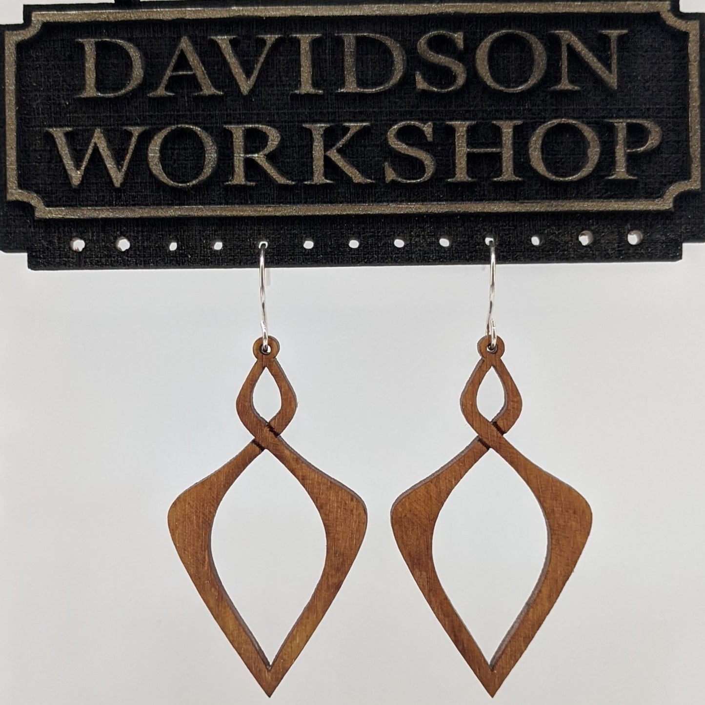 Pair of wooden earrings with silver stainless steel hooks. They are light natural finish elongated infinity symbols with sharp points. Made from birch wood hanging from a model Davidson Workshop sign against a white background. (Close up View)